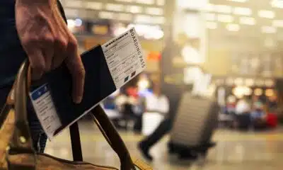 Imagem mostra uma mão segurando passaporte e passagem aérea em aeroporto.