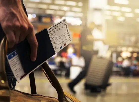 Imagem mostra uma mão segurando passaporte e passagem aérea em aeroporto.